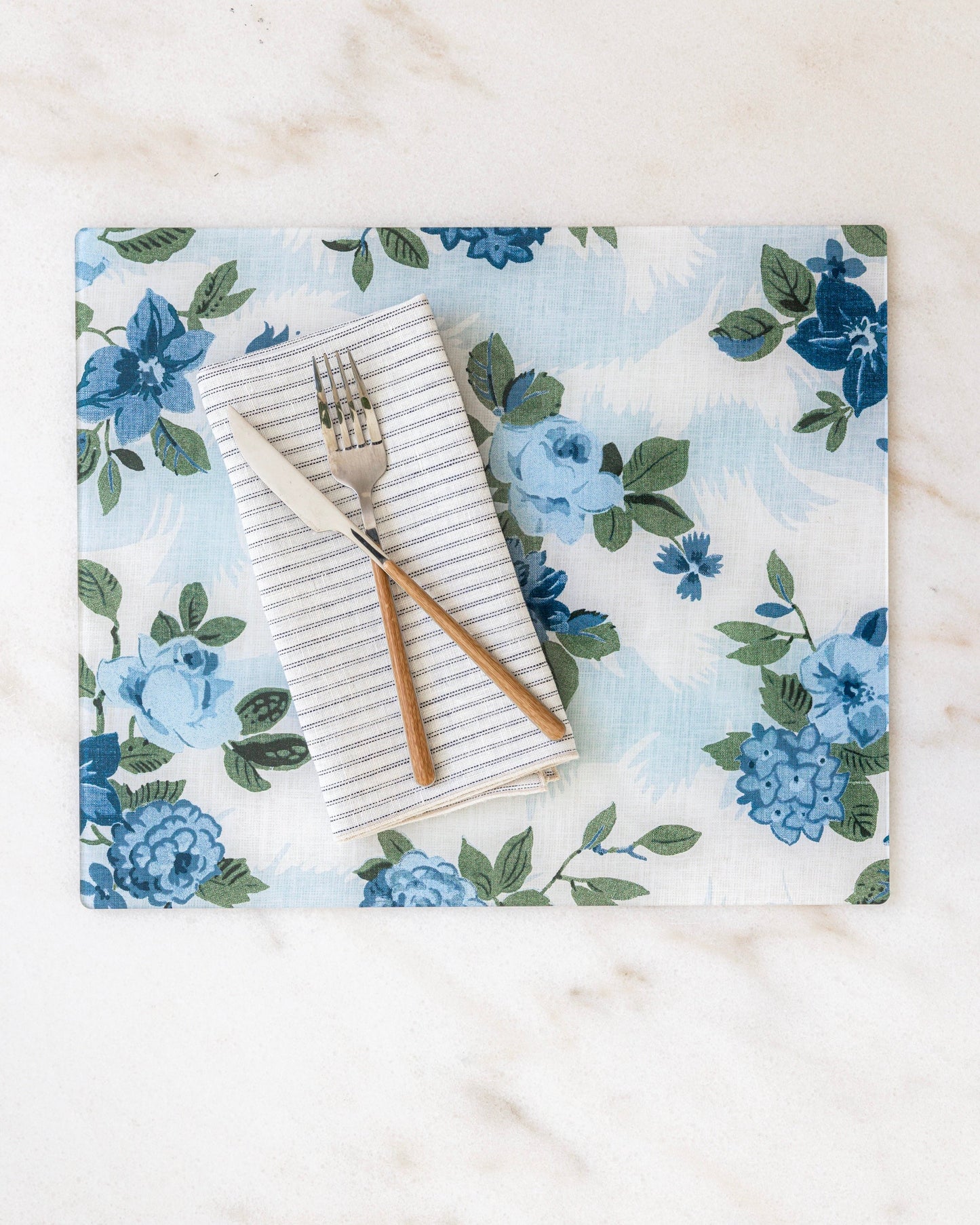 Floral placemat in shades of blue with a striped napkin and cutlery. The placemat is a collaboration between Proper Table and Nathan Turner Interiors. 