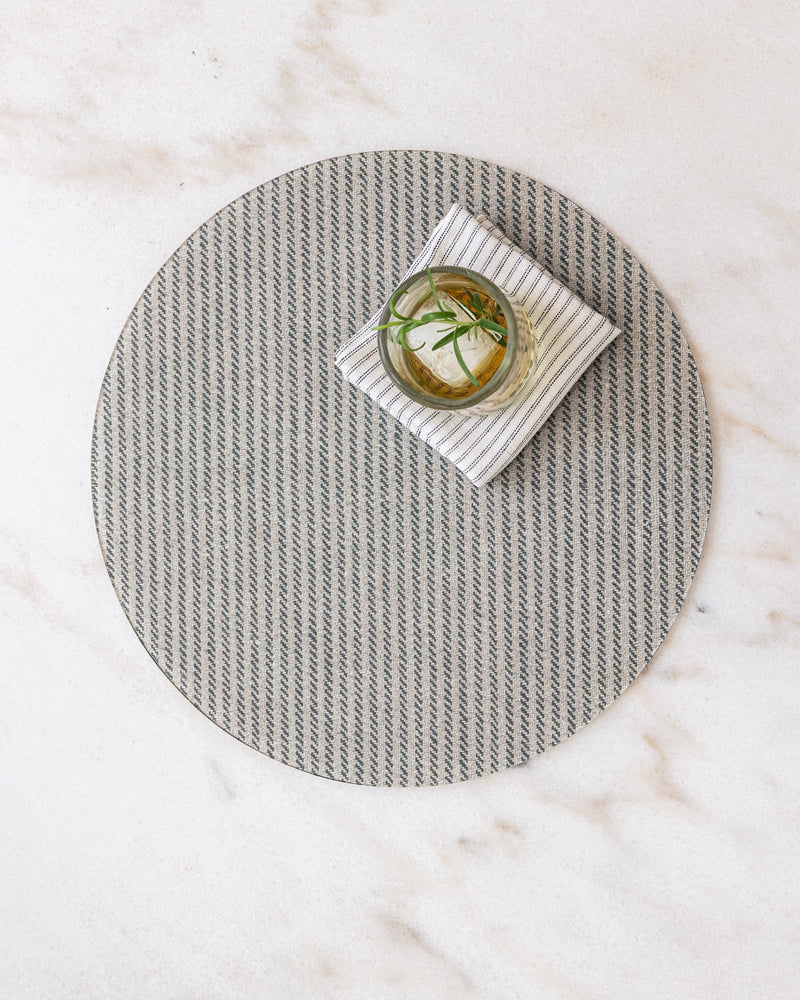 Round herringbone placemat in beige and gray on a marble surface with a small dish and a Proper Table linen ticking napkin.