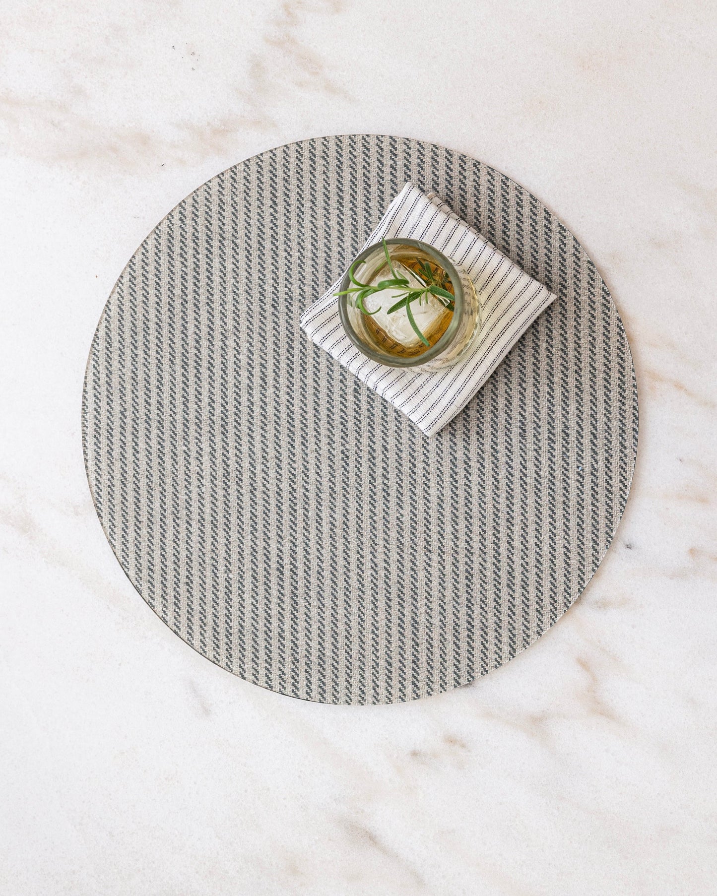 Round herringbone placemat in beige and gray on a marble surface with a small dish and a Proper Table linen ticking napkin.