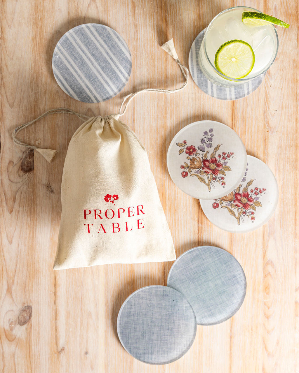 Proper Table coaster stack with a mix of Leah O'Connell x Proper Table floral coasters and striped and solid gray coasters on a  wooden surface with a 'Proper Table' gift bag.