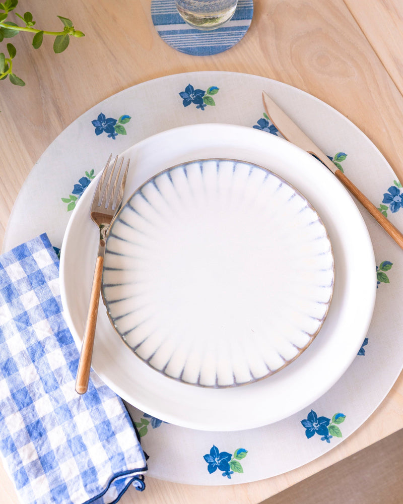 
                  
                    Blue and white round floral placemat on a table with striped coasters and gingham napkin.
                  
                