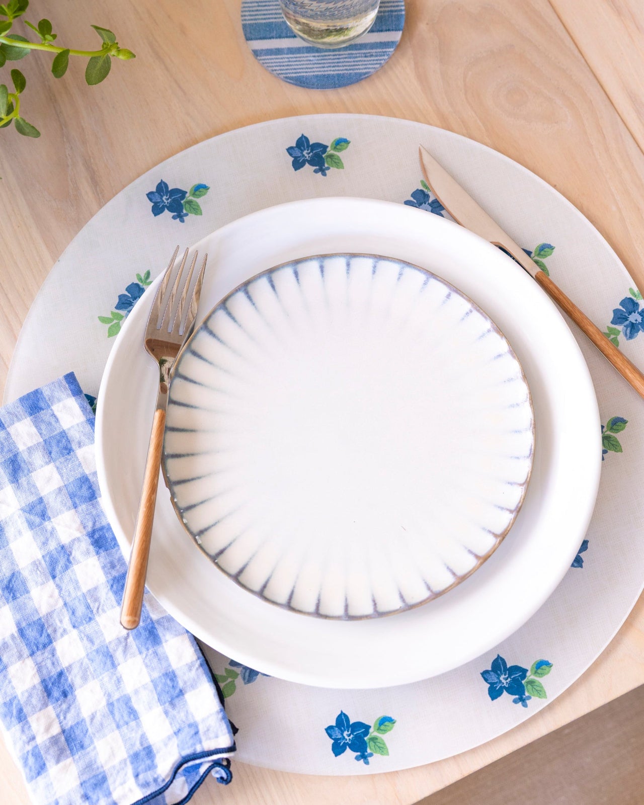 
                  
                    Blue and white round floral placemat on a table with striped coasters and gingham napkin.
                  
                