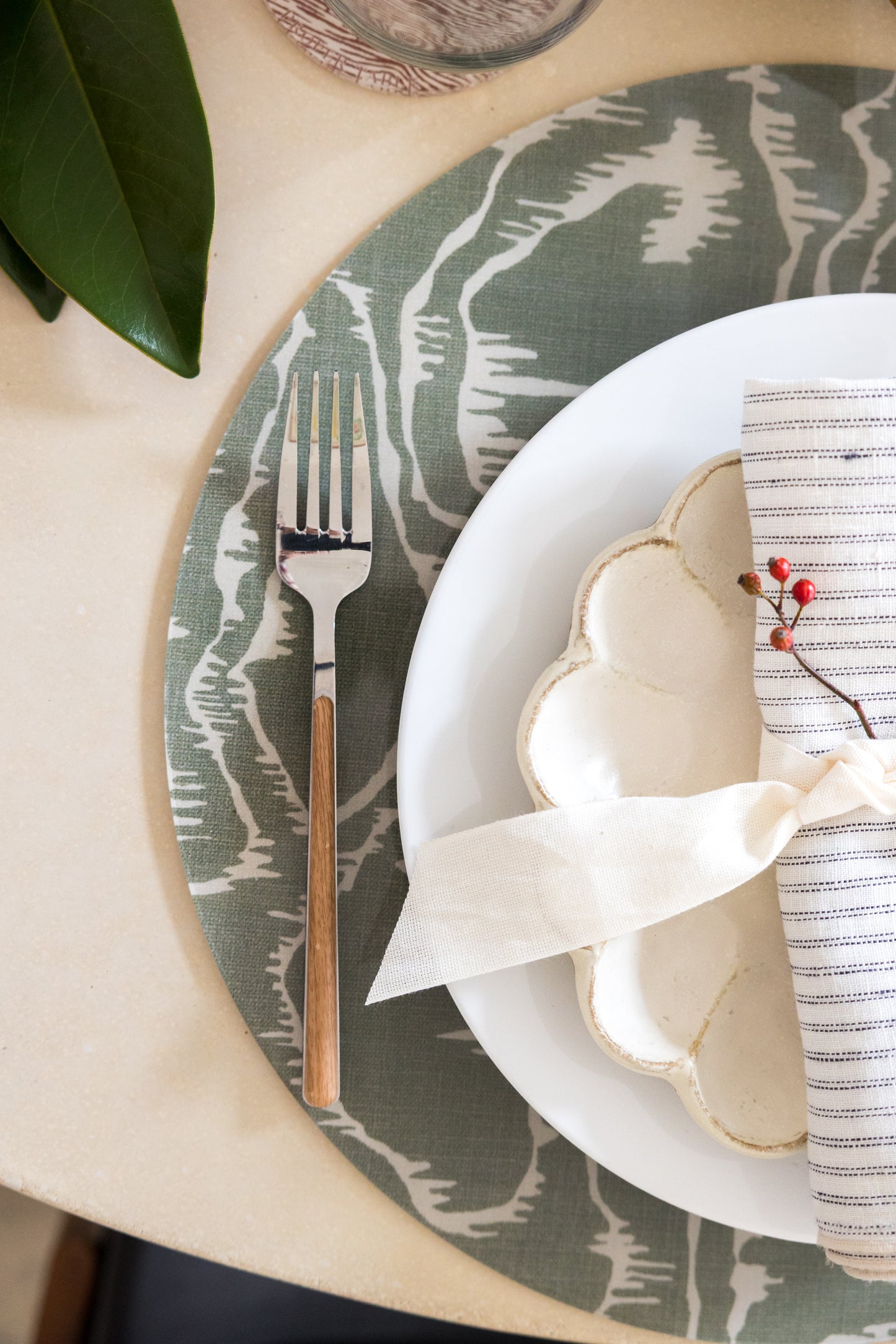 Round Tilda placemats from Proper Table x Leah O’Connell, styled in a simple holiday table setting with magnolia leaves, ribbon-tied napkins, and red berries. Placemats are green faux bois pattern.