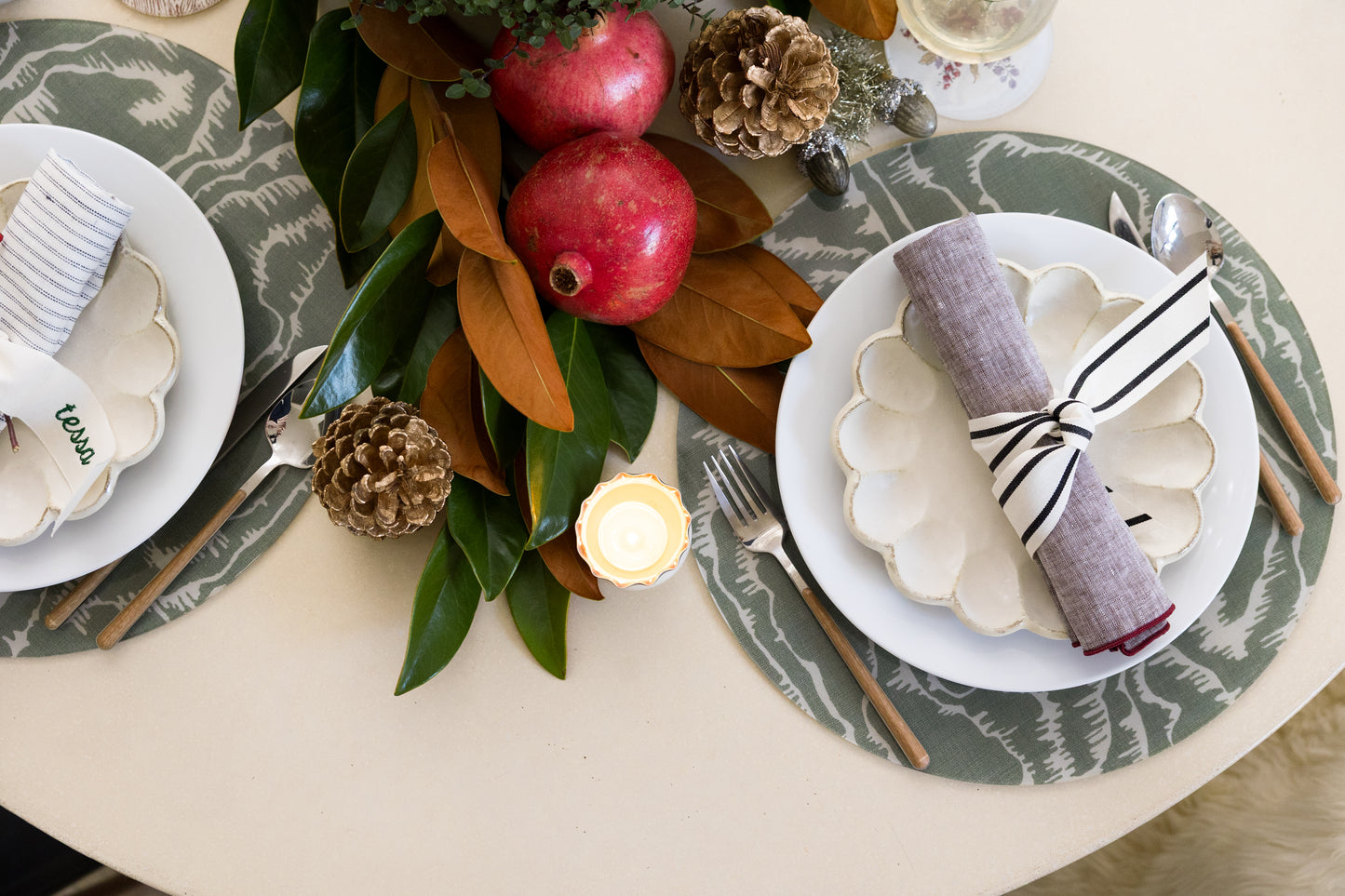 
                  
                    Tilda Round green placemats from Proper Table x Leah O’Connell in a festive holiday tablescape with magnolia leaves, pomegranates, and candles. Placemats are faux bois and acrylic 
                  
                