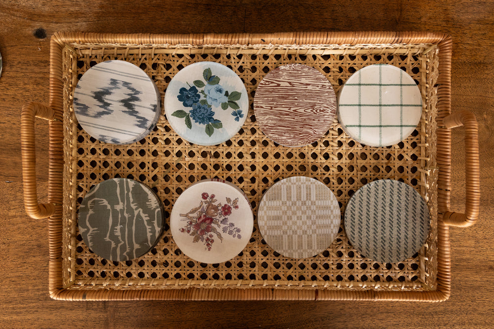 Sets of round acrylic coasters from Proper Table x Leah O’Connell in assorted patterns including ikat, floral, faux bois, and plaid, displayed in a woven tray.
