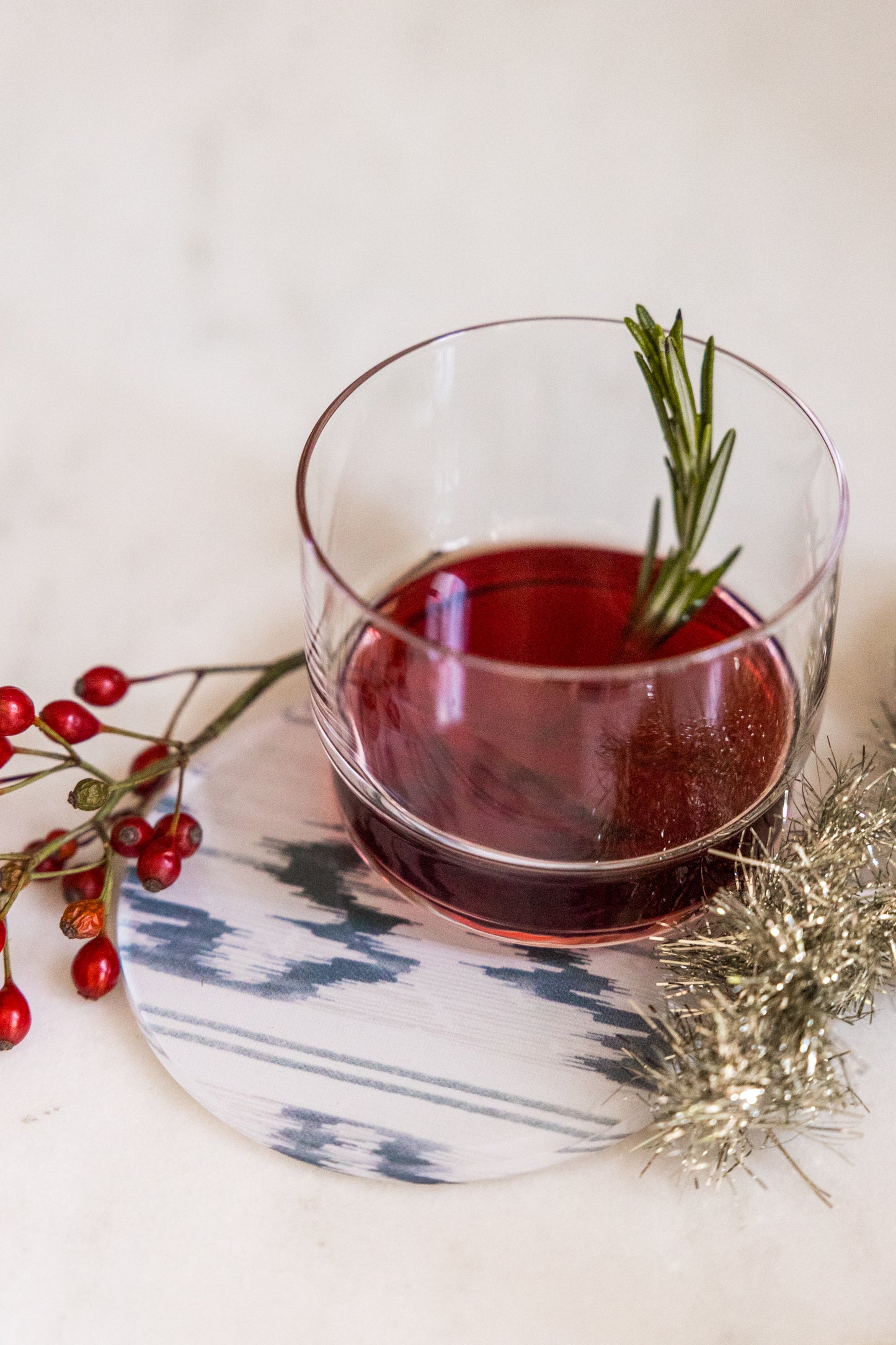 Round blue and white ikat acrylic coaster from Proper Table x Leah O’Connell, styled with a holiday cocktail garnished with rosemary and festive berries. Coaster is Isla ikat.