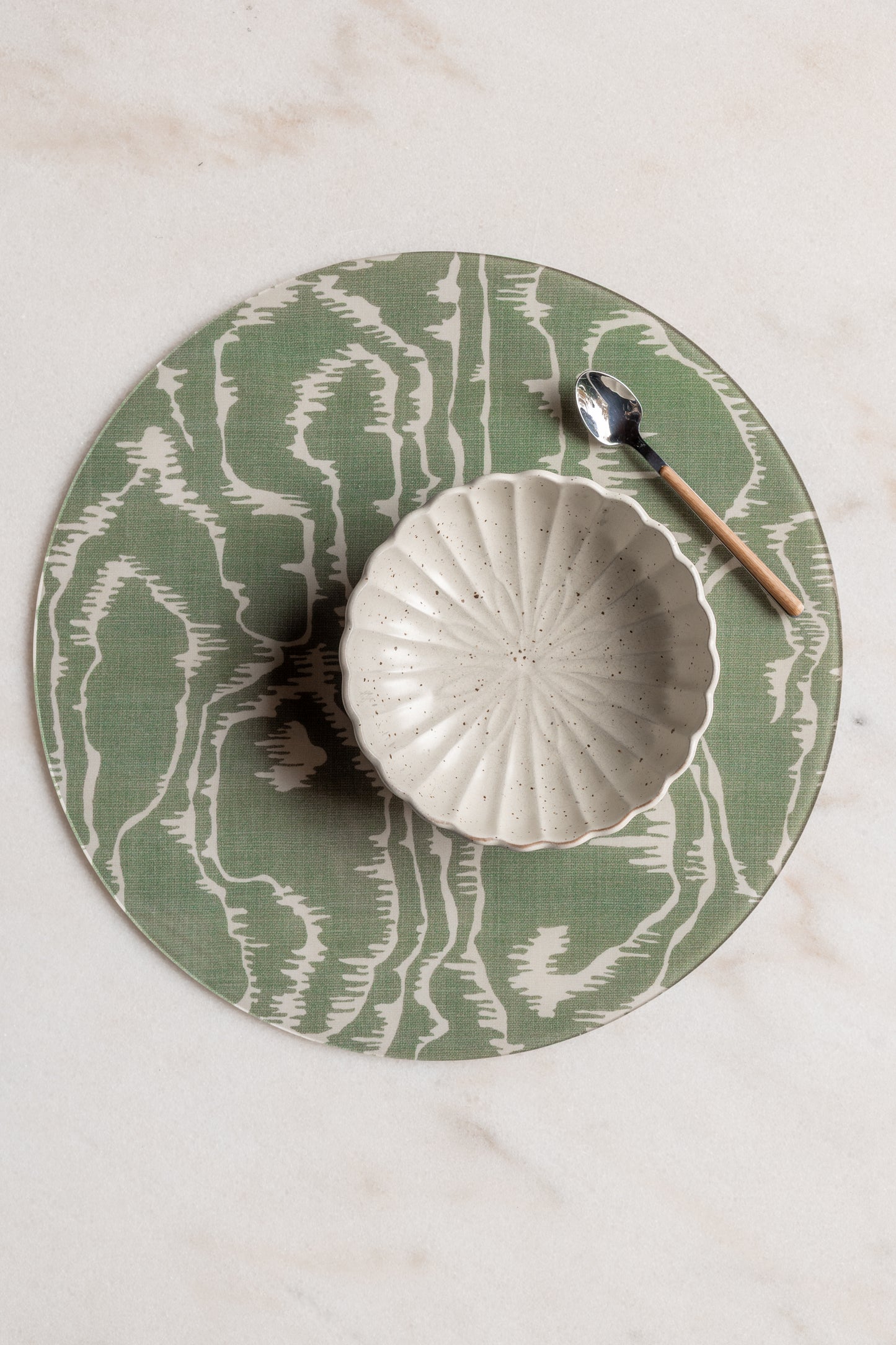 Round green and cream faux bois acrylic placemat from Proper Table x Leah O’Connell, styled with a scalloped ceramic bowl and wooden-handled spoon on marble.