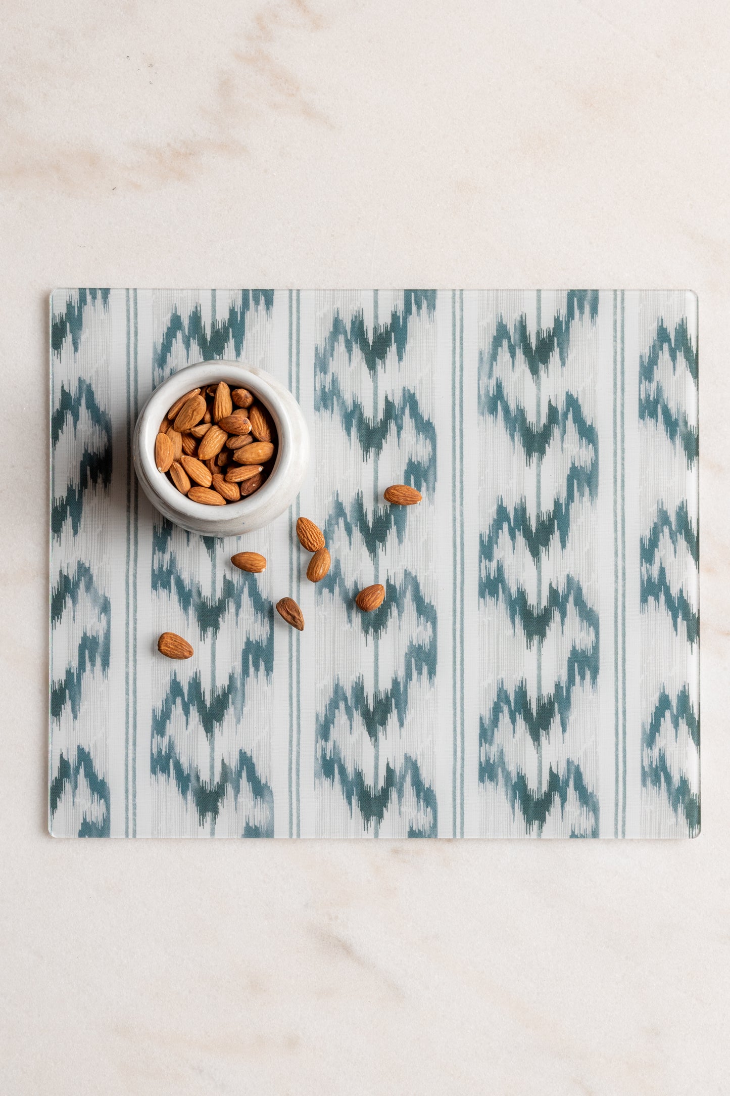 Rectangular blue and white ikat acrylic placemat from Proper Table x Leah O’Connell, styled on marble with almonds in a small white bowl. Placemat fabric is Isla from Leah O'Connell Textiles.